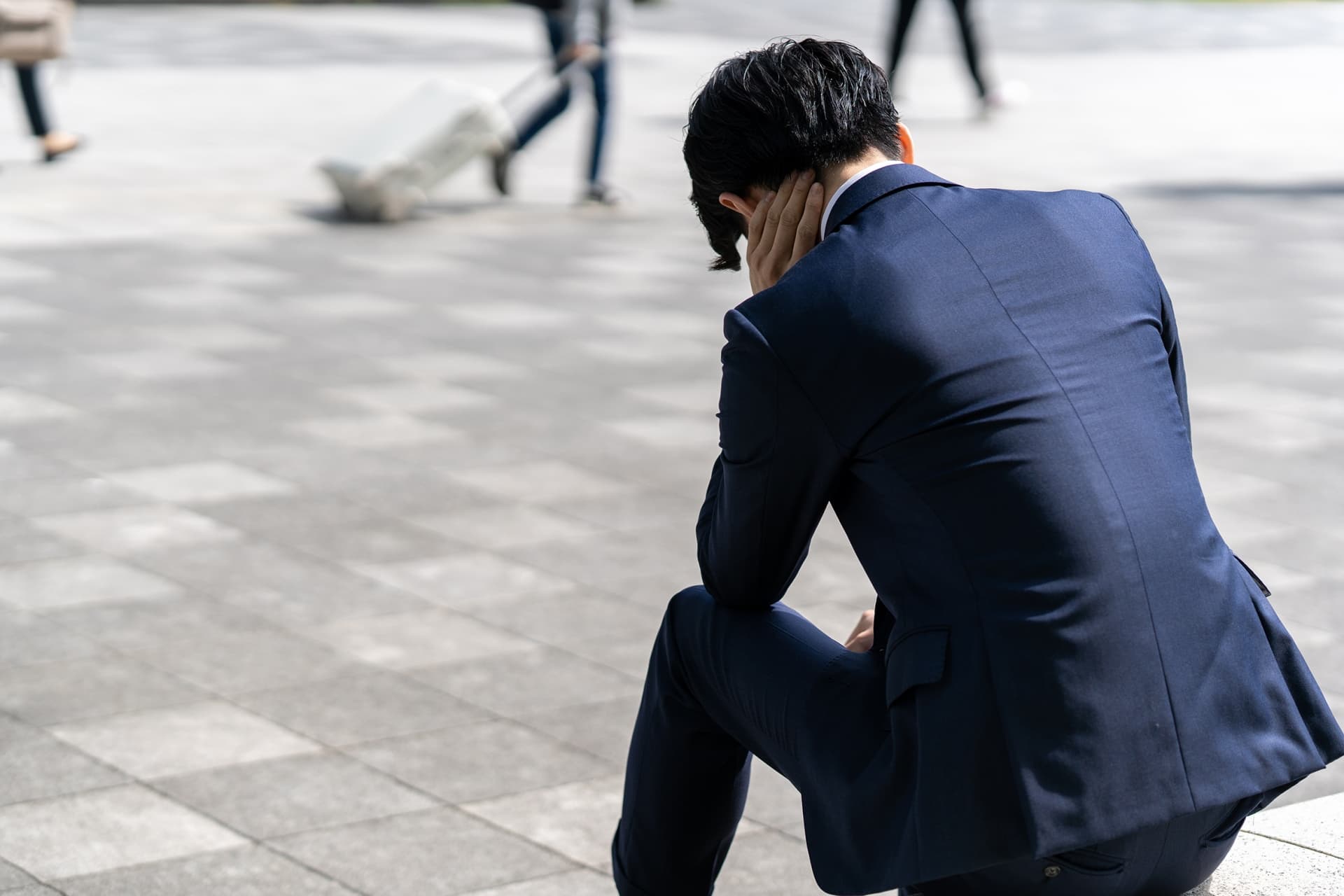 人通りのある広場で、ひとりうずくまるスーツ姿の男性の後ろ姿